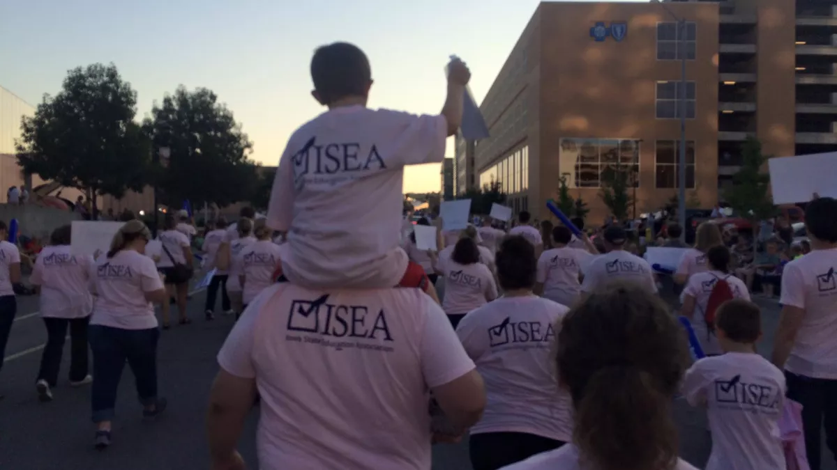 A picture from behind many ISEA members and supporters are marching. You can see their backs; they are in pink shirts with the blue ISEA logo. In the foreground, one person has a small child on their shoulders. The child is holding a sign. In the background, you can see the sun is beginning to set.