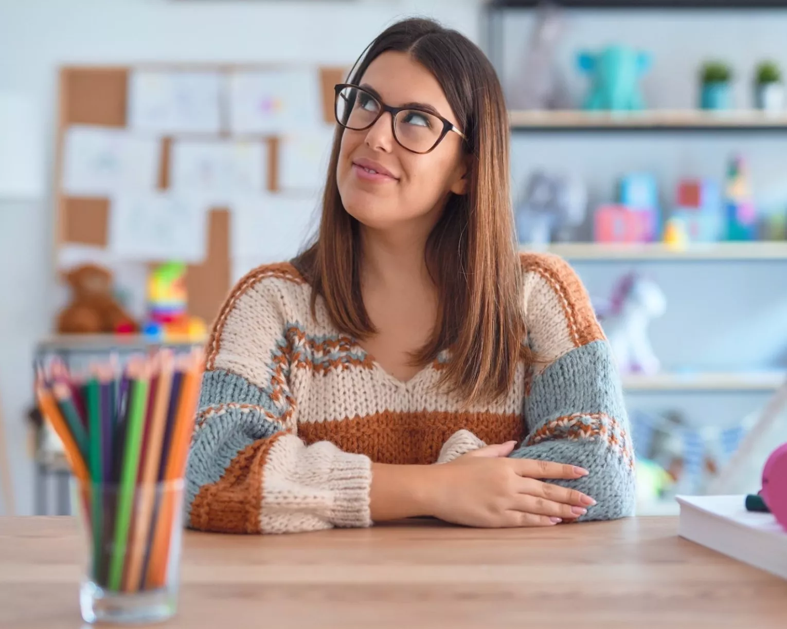 A white educator with long brown hair and black thick-rimmed glasses sits at her desk with a cup of colorful pencils in front her. She is smiling slightly and is looking up to her right like she has an idea.