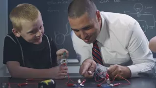 White main in white shirt and tie does science experiment with white blonde boy in a classroom.