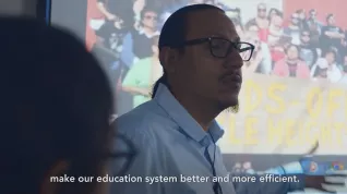 Teacher in front of projection with subtitle "make our educations system better and more efficient."