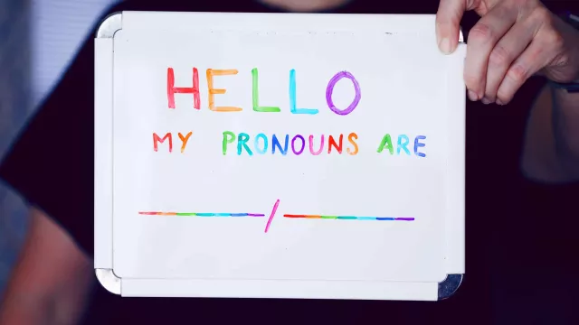 A woman holds a small whiteboard with a message inviting people to share their pronouns